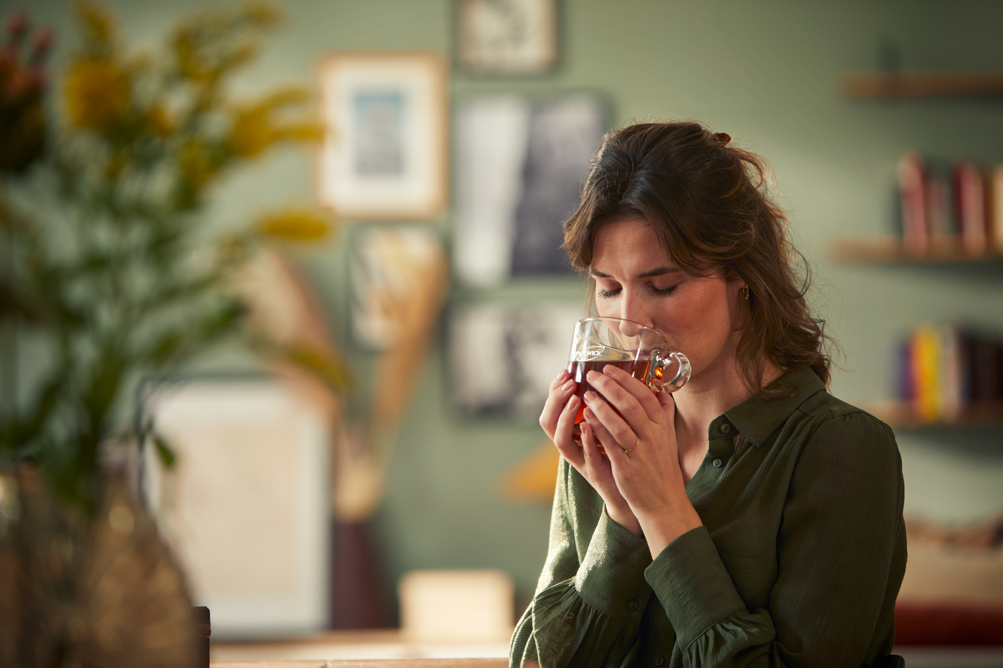Persoon houdt een glas Pickwick-thee vast voor de mond, met een groene muur, boekenplanken en gele bloemen op de achtergrond.