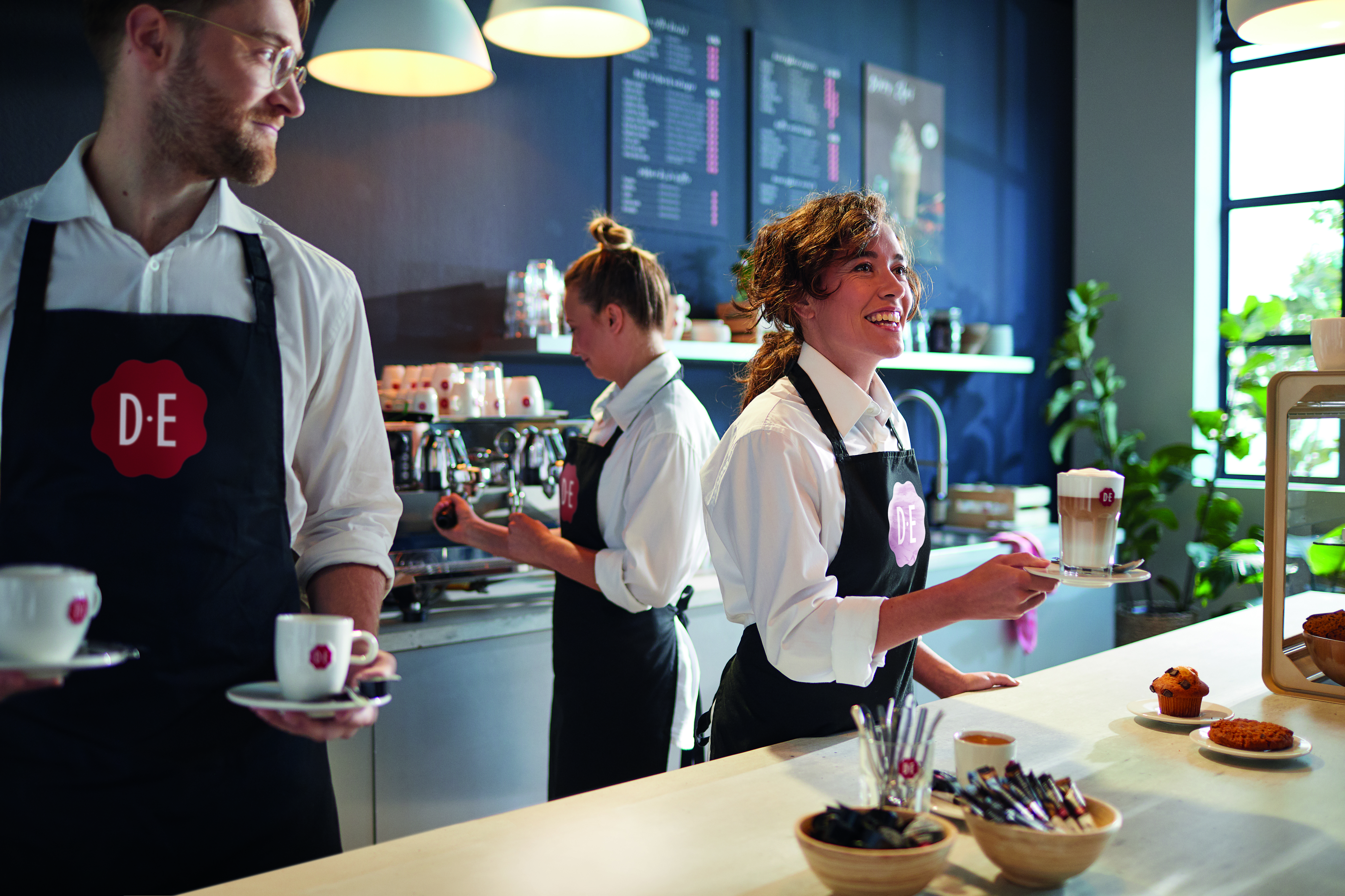 Drie barista’s werken in een koffiebar met D.E-logo, bezig met koffiezetten en serveren.